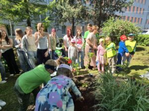 Wokół kawałka ziemi przygotowanego do nasadzenia roślin znajdują się uczniowie i dzieci. Wkładają kwiaty do ziemi i podlewają je.