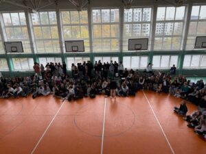 Zdjęcie przedstawia grupę osób zgromadzonych w szkolnej sali gimnastycznej z pomarańczową podłogą i wysokimi oknami, przez które wpada naturalne światło. Uczestnicy siedzą lub stoją wzdłuż obrzeża boiska, tworząc półokrąg. Przedstawiciele klas trzymają telefony i odpowiadają z członkami swojej grupy na pytania wyświetlane w kahoocie dotyczące książek, autorów i bohaterów literackich. Na twarzach widać emocje: skupienie, radość, czasem zaskoczenie po ogłoszeniu wyników.