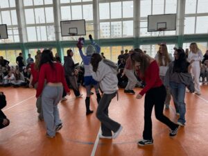 Zdjęcie przedstawia grupę uczniów uczestniczących w szkolnym wydarzeniu z okazji „Dnia Czytelnika”, zorganizowanym w sali gimnastycznej. Uczestnicy siedzą na podłodze, a na środku odbywa się scenka promująca książkę. Centralnym punktem jest grupa osób w przebraniach: niektórzy mają opaski z uszami, ogony lub fantazyjne stroje, co nadaje scence charakter telewizyjnego programu dla dzieci lub młodzieży. Uczniowie odgrywają humorystyczną sytuację, która ma na celu zachęcenie widzów do sięgnięcia po książkę. W tle siedzą uczniowie i obserwują przebieg wydarzenia.