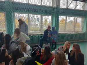 Zdjęcie przedstawia grupę osób zgromadzonych w sali gimnastycznej, z zieloną podłogą i dużymi oknami zabezpieczonymi siatką. Większość uczestników siedzi na podłodze w luźnej, towarzyskiej atmosferze. Na środku, na krześle siedzi dyrektor szkoły, a obok niego kuca uczennica przebrana za postać literacką: ma niebieski strój i pomalowaną twarz. Pozostali uczestnicy ubrani są swobodnie, niektórzy mają opaski z uszami myszy. Wszyscy wyglądają na zaangażowanych i zadowolonych ze szkolnego wydarzenia pt.” Dzień Czytelnika”.