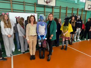 Zdjęcie przedstawia grupę osób zgromadzonych w szkolnej sali gimnastycznej z pomarańczowo-zieloną podłogą. W tle widoczne są drabinki gimnastyczne, kosze do koszykówki oraz tablica. Na środku zdjęcia stoją dwie nauczycielki ubrane w codzienne stroje. Towarzyszy im osoba przebrana za postać literacką, Pana Bobińskiego z książki „Karolina”. Ma pomalowaną na niebiesko twarz i ciało, a w ręce siatkę z burakami. W tle stoi grupa uczniów przebranych za różne postacie literackie.