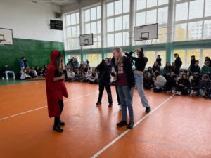 Zdjęcie przedstawia grupę osób zgromadzonych w szkolnej sali gimnastycznej z pomarańczową podłogą. Na środku stoi czterech uczniów. Po lewej znajduje się dziewczyna, przebrana za Czerwonego Kapturka, a po prawej chłopak z długimi włosami, z mikrofonem w ręce. Za nimi stoi dwójka uczniów z rękami w górze, udających drzewa. W tle siedzą na podłodze uczniowie.