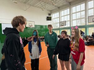 Zdjęcie przedstawia grupę osób zgromadzonych w szkolnej sali gimnastycznej z pomarańczową podłogą. Na środku stoi pięcioro uczniów, którzy biorą udział w konkurencji: kalambury literackie. Po lewej stronie stoi uczeń z kartką w ręku, a naprzeciwko niego, od prawej: uczennica przebrana za mysz, dziewczynka z namalowaną blizną na twarzy, chłopak w zielonej bluzie z mikrofonem w ręku i dziewczyna z twarzą pomalowaną na niebiesko. W tle siedzą na podłodze uczniowie.