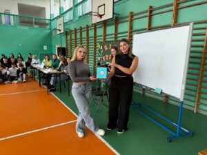 Zdjęcie przedstawia trzy uczennice w szkolnej sali gimnastycznej z pomarańczową podłogą. Jedna uczennica ubrana na czarno wręcza drugiej niebieską torebkę prezentową, jako nagrodę w konkursie: kahoot literacki. Po prawej stronie znajduje się biała tablica, w tle siedzi jury oraz uczniowie.