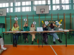 Zdjęcie przedstawia pięcioosobowe jury, siedzące na krzesłach za stołem w szkolnej sali gimnastycznej z pomarańczową-zieloną podłogą. W tle znajduje się tablica z gazetką promująca czytelnictwo oraz drabinki. Jury trzyma w rękach tabliczki z przyznaną punktacją za zadanie w turnieju klas. Od lewej członkowie jury pokazują: trzy punkty, trzy punkty, dwa punkty, dwa punkty i dwa punkty.