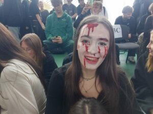 Zdjęcie przedstawia uśmiechniętą uczennicę z twarzą pomalowaną na biało, z czerwonymi plamami, imitującymi krew. Dookoła niej siedzą na podłodze uczniowie, dwóch chłopców za nią trzyma w rękach telefony i grają w kahoota literackiego.