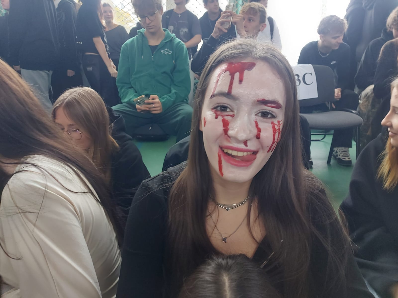 Zdjęcie przedstawia uśmiechniętą uczennicę z twarzą pomalowaną na biało, z czerwonymi plamami, imitującymi krew. Dookoła niej siedzą na podłodze uczniowie, dwóch chłopców za nią trzyma w rękach telefony i grają w kahoota literackiego.