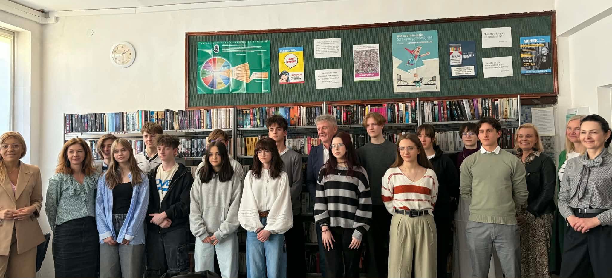 Zdjęcie przedstawia dużą grupę ludzi: uczniów, nauczycieli, gości, stojacych w dwóch rzędach. W środku stoi dyrketor szkoły, P. Radosław Jankowski. W tle widać półki z książkami. U góry na ścianie po lewej stronie wisi zegar, a po prawej tablica z plaktami.