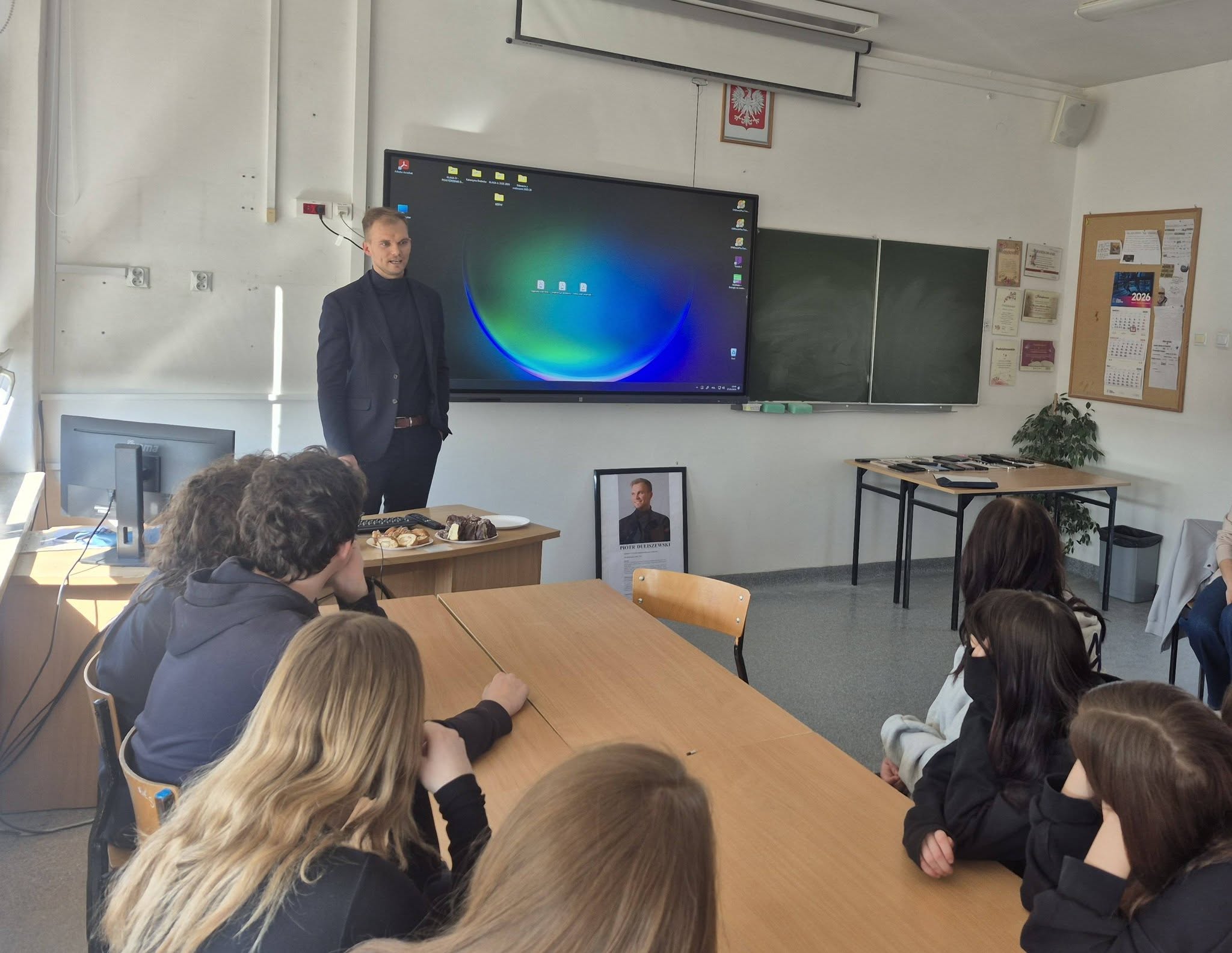 Absolwent szkoły stoi za biurkiem i rozmawia z uczniami. Na zdjęciu widocznych jest kilku uczniów siedzących przy stolikach i odwróconych plecami. Patrzą na absolwenta. Ten ubrany jest w garnitur. Za jego plecami znajduje się tablica multimedialna i zwykła. Rozmowa toczy się w sali szkolnej.