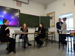 Na zdjęciu widać 5 osób, wszystkie są przebrane. Trzy osoby siedzą przy stole, za plecami mają tablicę (multimedialną i kredową), dwie stoją przy drzwiach. Wszyscy ubrani są w stroje z epoki międzywojnia. Odgrywają scenkę, w której sąd (w rolę sędziny wciela się uczennica) ma orzec winę lub niewinność artysty awangardowego.