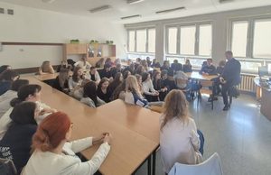 Absolwent siedzi z przodu sali na krawędzi stołu. Rozmawia z uczniami. Uczniowie i nauczyciele patrzą w jego kierunku. Stoliki ustawione są w podkówkę, pośrodku sali stoją ławeczki zajęte przez młodzież.