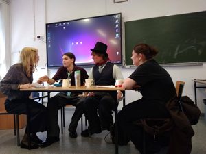 Na zdjęciu widać 4 osoby ubrane w epokowe stroje. Siedzą przy stoliku i wcielają się w rolę artystów międzywojnia. Do konfliktu dochodzi między surrealistką, futurystką, dadaistą i katastrofistą.