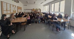 Na zdjęciu widoczni są nauczyciele i młodzież uczestnicząca w spotkaniu z absolwentem. Siedzą przy stolikach ustawionych w podkówkę. Pośrodku znajdują się ławeczki – także zajęte przez uczniów. Twarze wszystkich zwrócone są w kierunku absolwenta, którego na zdjęciu nie widać.