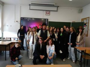 Zdjęcie to zostało zrobione po odegraniu scenek projektowych i ma charakter pamiątkowy. Pozują na nim osoby z klasy 3b z polonistą. Część osób stoi, kilka kuca. Zdjęcie zostało zrobione w sali szkolnej. Widać elementy wyposażenia klasy, np. tablicę.