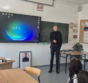 Ubrany w garnitur absolwent stoi pośrodku sali i rozmawia z uczniami. Za plecami ma tablicę multimedialną i zwykłą. Jego gesty świadczą, że zadaje pytania. Na podłodze stoi antyrama z podobizną absolwenta.