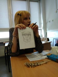 Uczennica siedzi przy stoliku i trzyma kartkę, na której narysowany jest wizerunek artysty awangardowego. Ubrana jest w strój z epoki, całości dopełniają drobne elementy dekoracyjne.