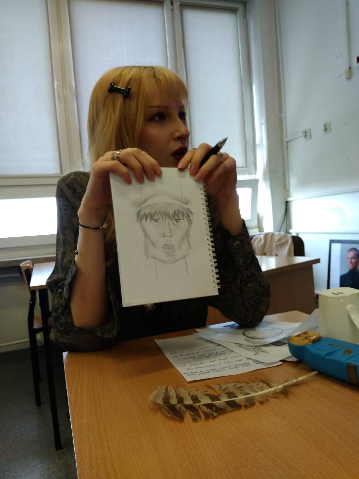 Uczennica siedzi przy stoliku i trzyma kartkę, na której narysowany jest wizerunek artysty awangardowego. Ubrana jest w strój z epoki, całości dopełniają drobne elementy dekoracyjne.